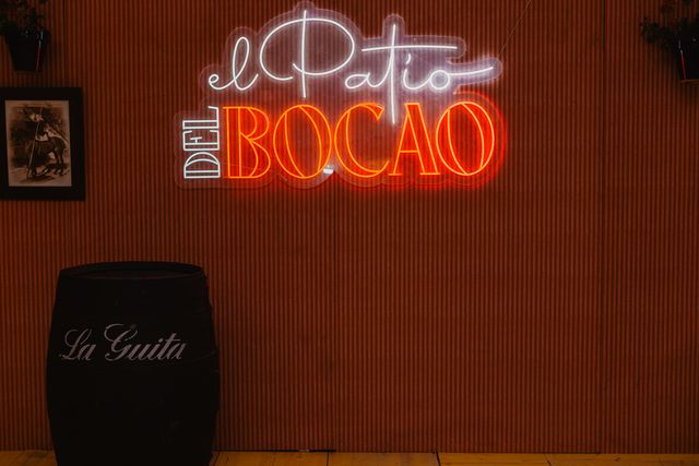 Neón «El Patio del Bocao» sobre la pared de rayas, con una barrica de La Guita